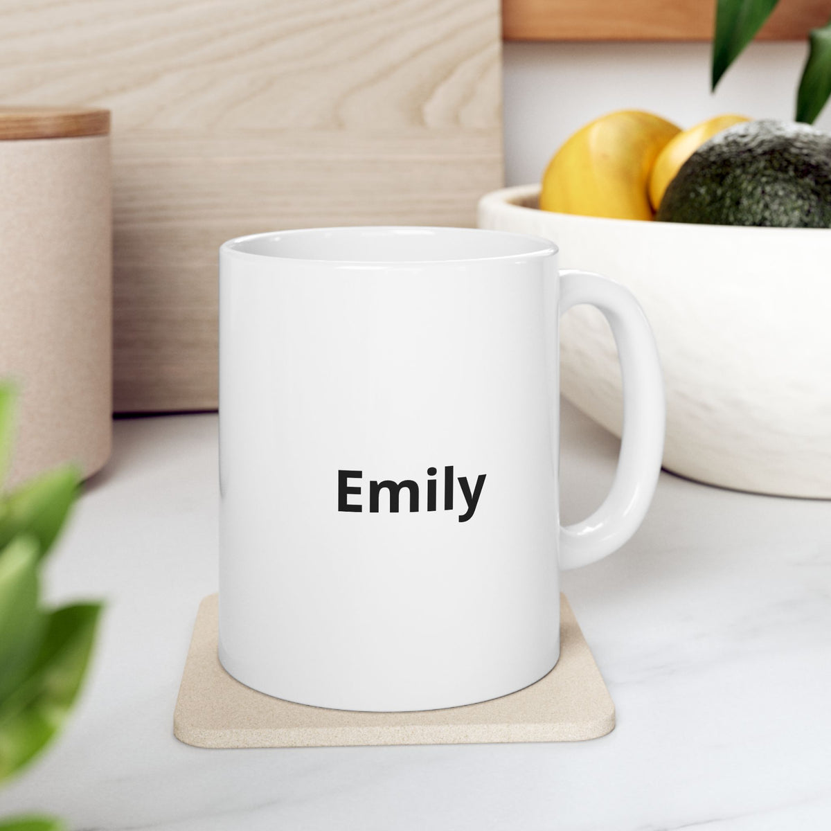 White mug personalized with Emily on a coaster beside a fruit bowl in a kitchen.