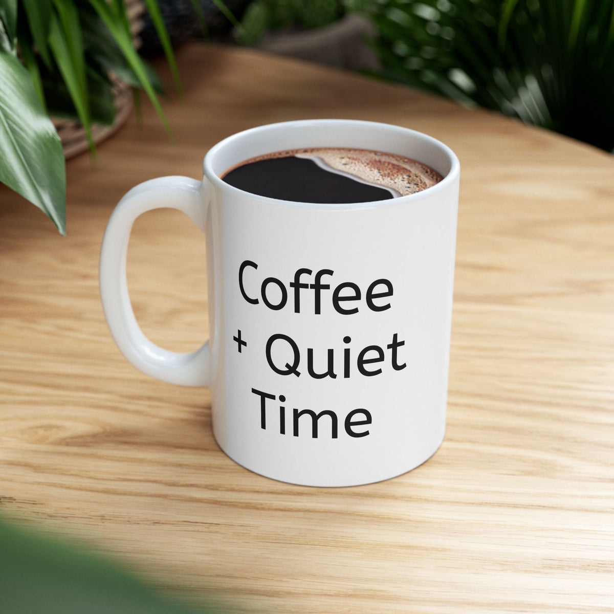 Coffee + Quiet Time mug filled with coffee on a light wooden table beside green leaves.