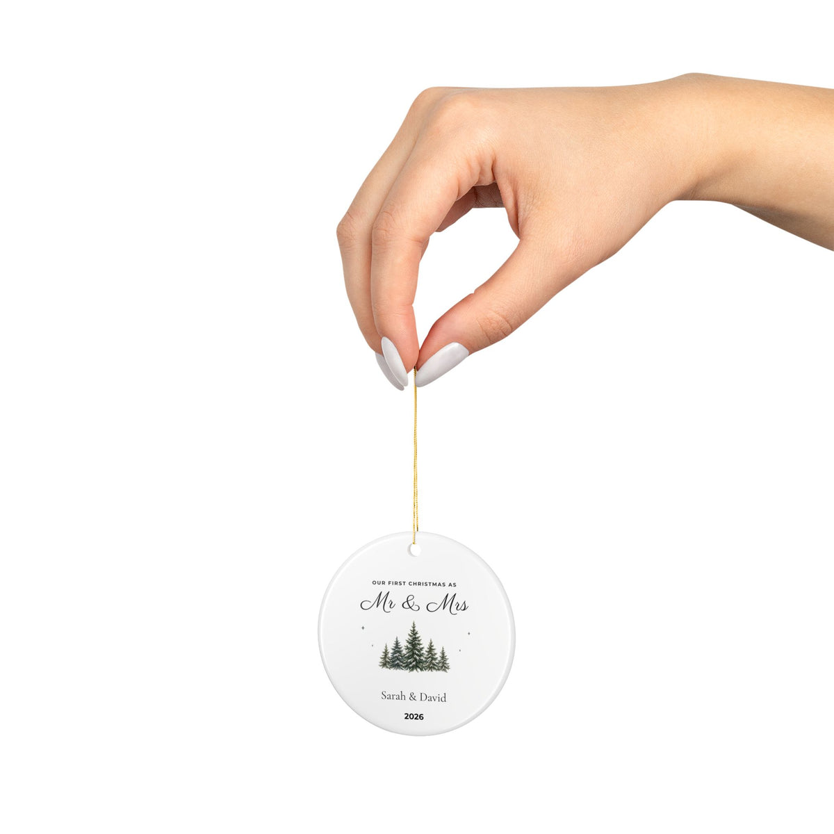 Hand holding the personalized First Christmas Mr & Mrs ceramic ornament by its gold hanging cord, showing evergreen trees, example names Sarah & David and year 2026.
