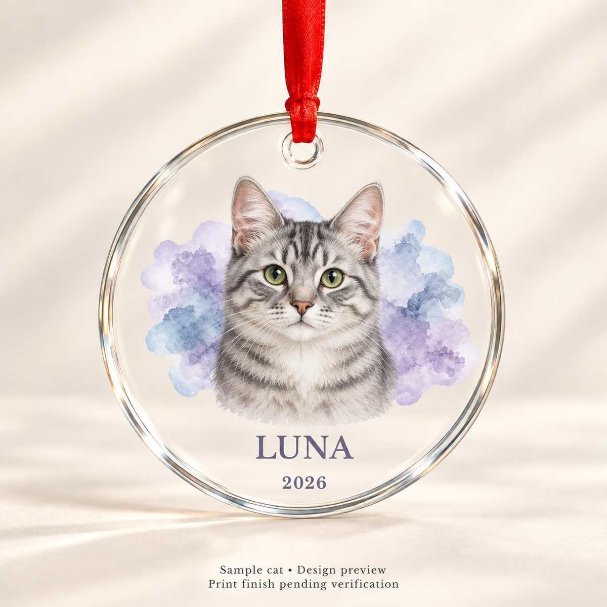 Design preview of Luna silver tabby cat keepsake glass ornament with lavender-blue watercolor, name, 2026 and red satin ribbon. Print finish pending verification.